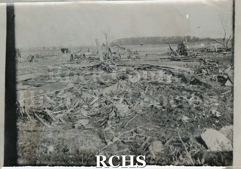 Black and white photograph of damage from tornado of 1911. Debris is spread throughout the field. A tractor is visible among the debrfis.