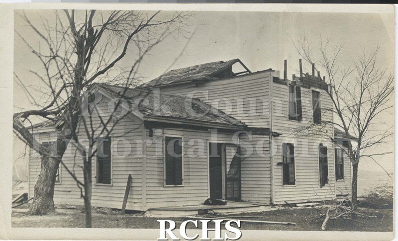 Black and white photograph of the tornado of 1911. Shows a house damaged by a tornado. The roof is off and windows are damaged.