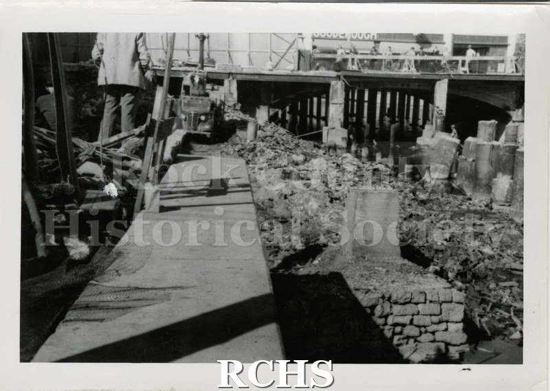 Black and white photograph of the foundations of a building. There is a sidewalk on the left and a man standing in the debris.