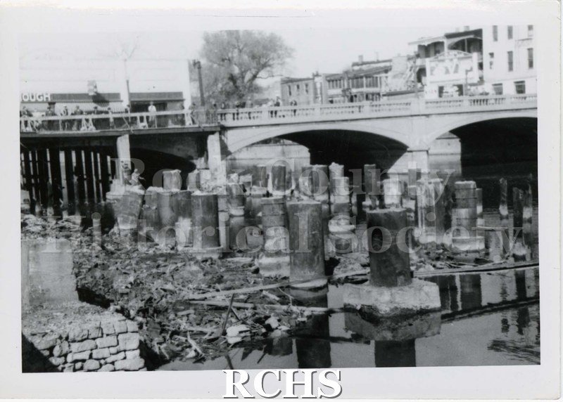 Black and white photograph of building pilings in the river in front of the bridge. Buildings are visible at top of photograph.
