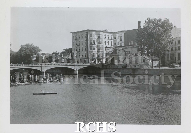 Black and white photograph of building pilings in the river in front of the bridge. Buildings are visible at top of photograph.