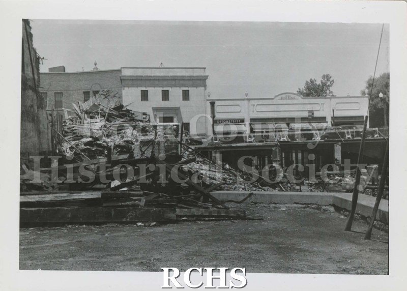 Black and white photograph of building ruins after 1942 fire. Debris is piled up on shore of the river. There are buildings visible in the background.