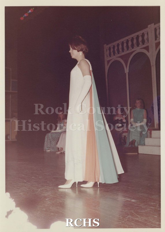 Colored photograph of Suzi Soeder, Contestant for Miss Janesville 1968, wearing a white formal with a colored train and long white gloves.