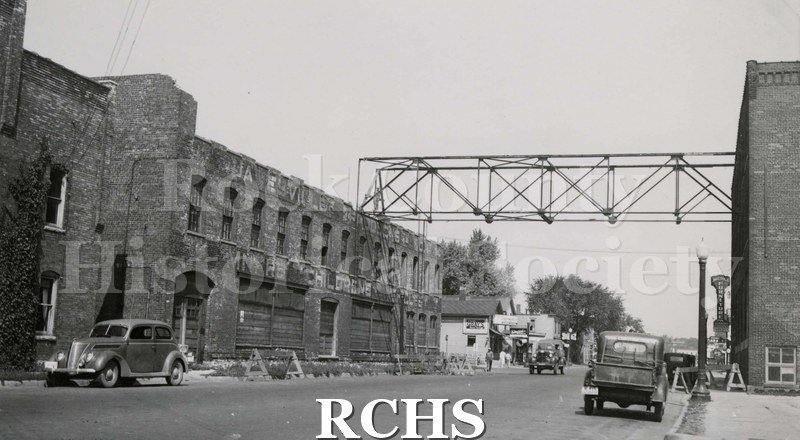 Black and white photograph of Janesville Products, Janesville, WI. Pictured is a two story building connected by a walkway to another building on the right.