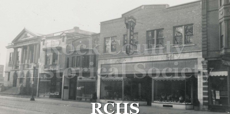 Black and white photograph of Main Street, Janesville, WI, showing Janesville Floral Company on the right and the public library on the left.
