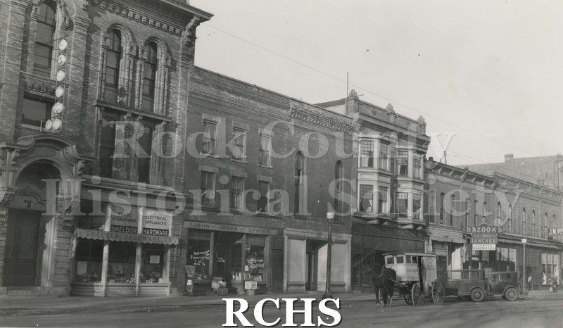 Black and white photograph of Main Street, Janesville, WI, showing stores: Sheldon Hardware; Razook's Lunches; and J. M. Bostwicks.