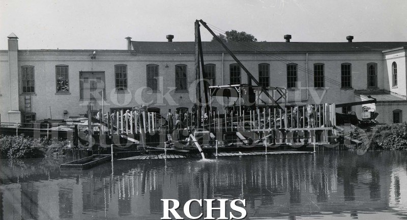 Black and white photograph of Rock River Woolen Mills 1943. Pictured is a building on the river with workmen working on the outside and machinery in front.