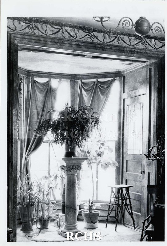 Black and white photograph of the interior of house at 317 Madison St., showing the window with many plants on the floor and on a pedestal.