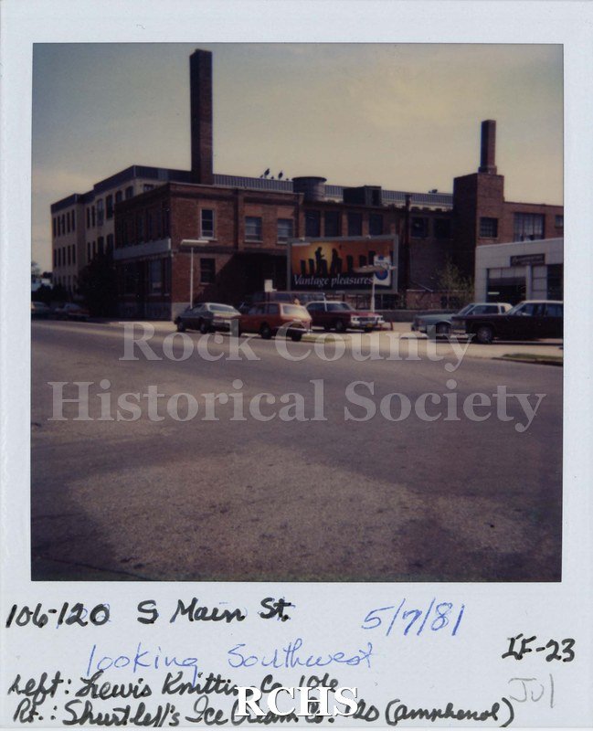 Black and white photograph of 106-120 S. Main Street, a two story brick building with a billboard in front and cars parked on the street.