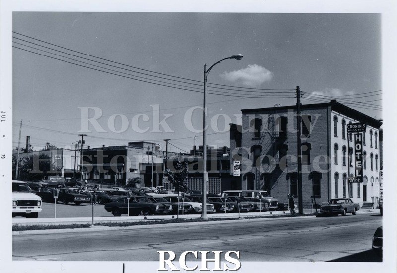 Black and white photograph of several buildings on Main Street. Cronin's Hotel is on the right with many cars in the parking lot beside it.