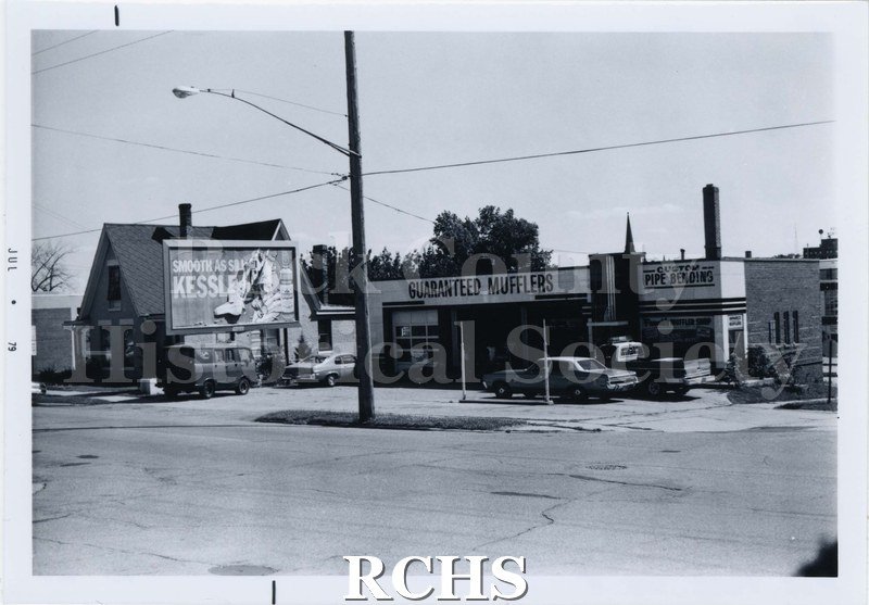 Black and white photograph of 217 and 221 Parker Drive, North. Shows a building with signs for "Guaranteed Mufflers" and "Custom Pipe Bending".