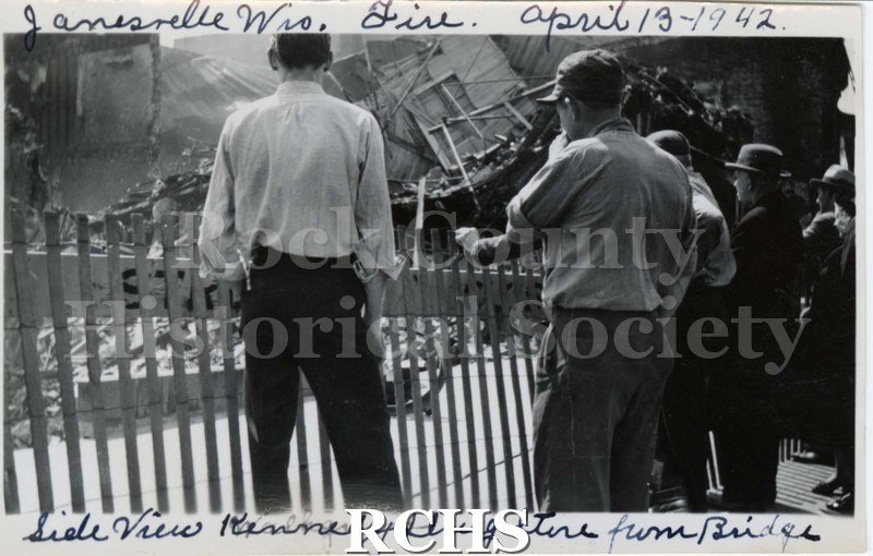 Black and white photograph of Janesville Wis.Fire April 13, 1942. Shows spectators standing near a fence. On bottom: "Side View Kennedy Drug Store from Bridge".