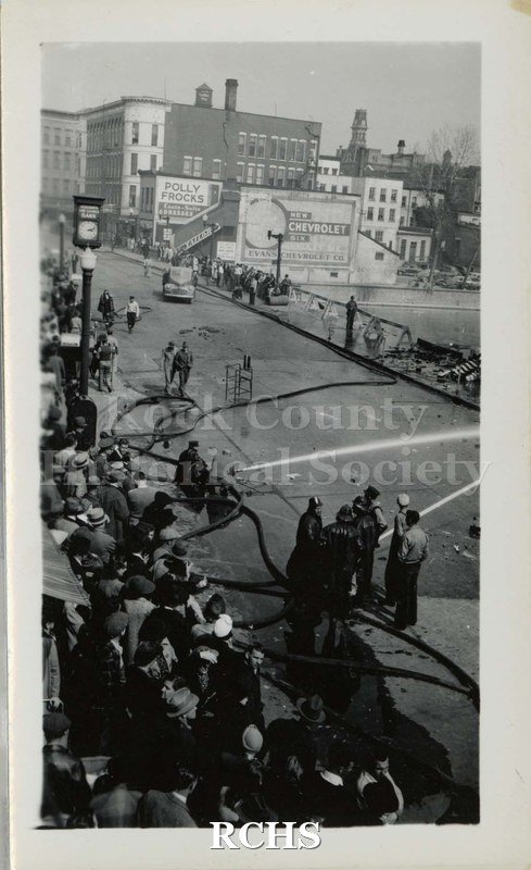 Black and white photograph of a crowd of people watching the firemen play their hoses on a burning building (not visible).