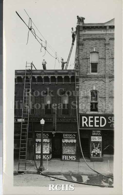 SPhotograph. Black and white photograph of several men on top of the drug store building. One is on a ladder and one is on the roof of the Reed building.