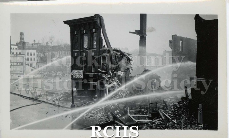 Black and white photograph of streams of water being poured on demolished buildings from a fire. Sign on building on the left reads; "Kennedy Drugs".