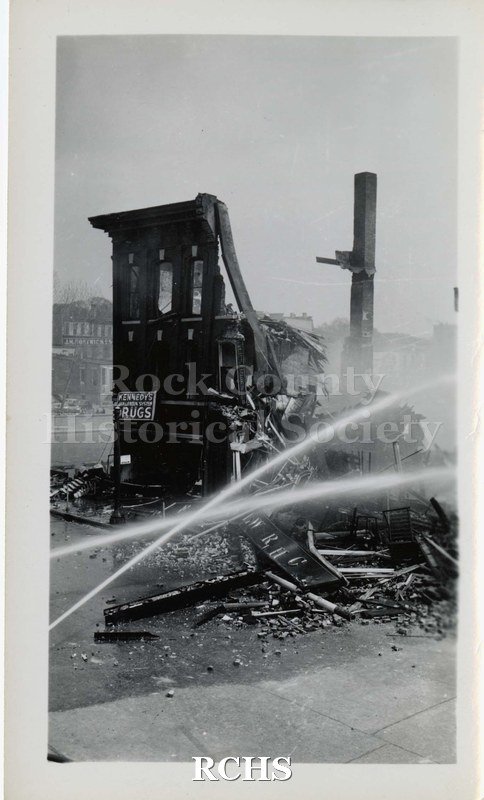Black and white photograph of streams of water being poured on demolished buildings from a fire. Sign on building on the left reads; "Kennedy Drugs".