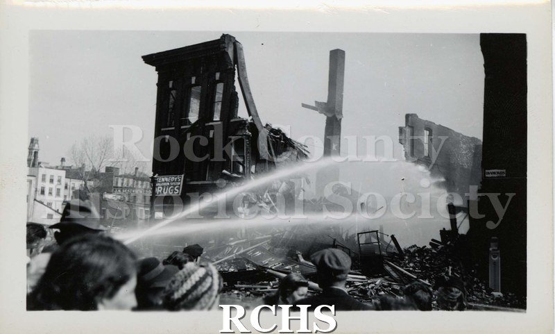 Black and white photograph of streams of water being poured on demolished builidngs from a fire. Sign on building on the left reads; "Kennedy Drugs".