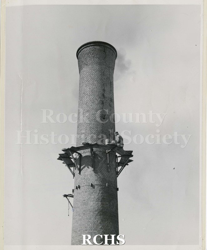 Black and white photograph of Carl Stanley seated on scaffolding on what is likely smokestack of Janesville High School. Appears to be tuck pointing.