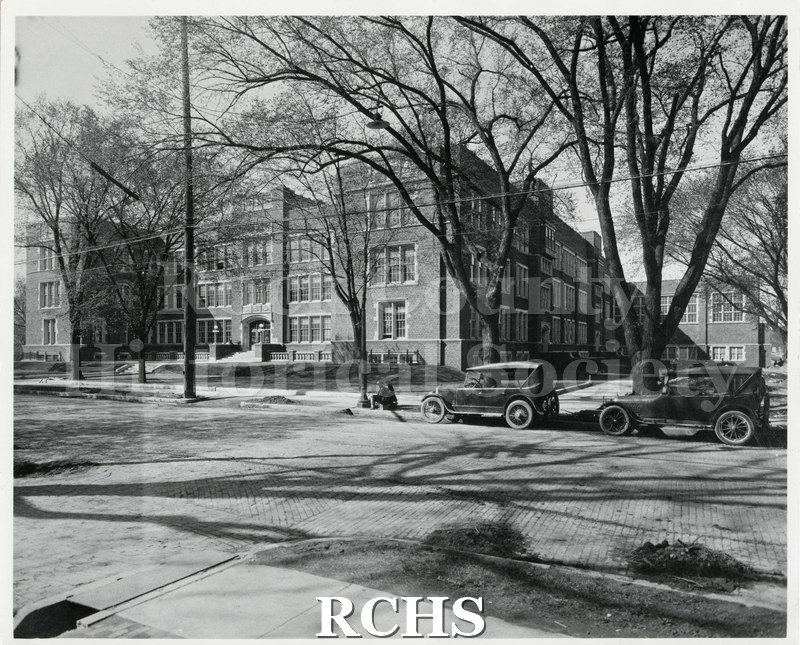 Black and white photograph of Janesville High school, taken from across Main Street. Two figures are seated alongside the curb in front of two vehicles.