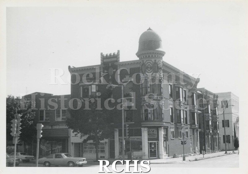 Black and white photograph of Kent block, a building in downtown Janesville. Shows a three story brick building with a rounded front.