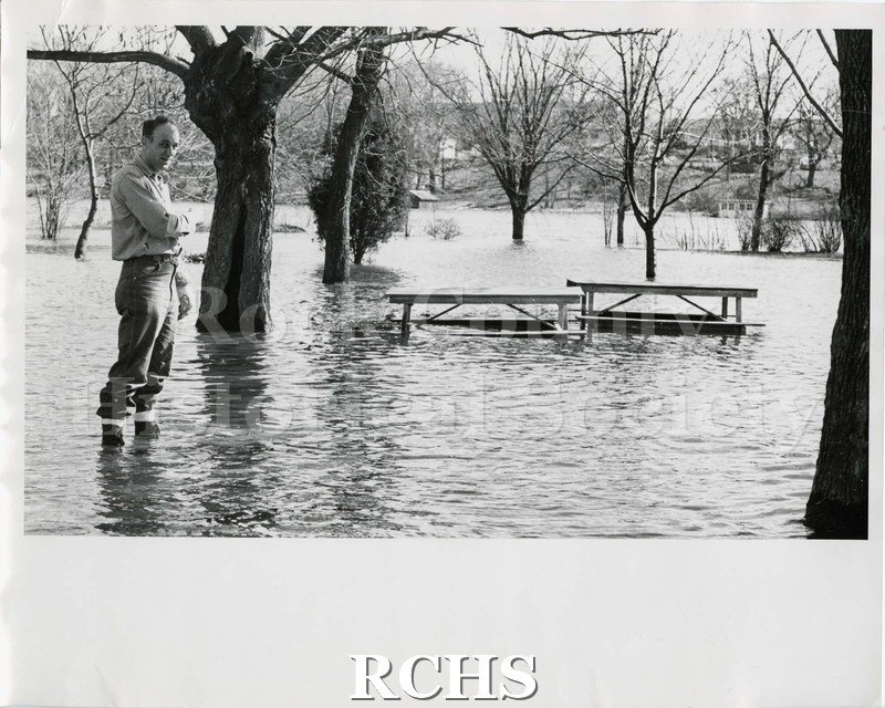 Black and white photograph of flooding of Rock River. Shows a man standing in water in a park. There are two picnic tables and several trees visible.