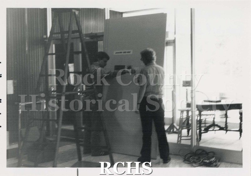 Black and white photograph of the Janesville Public Library Centennial Exhibit Installation. Shows two people working on a door.