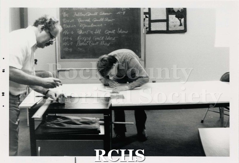 Black and white photograph of the Janesville Public Library Centennial Exhibit Installation. Shows two people working on a door.