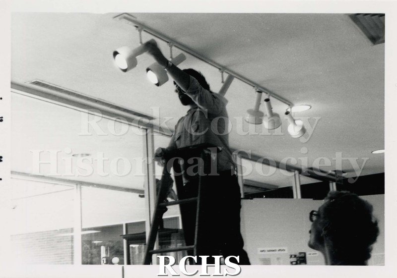 Black and white photograph of the Janesville Public Library Centennial Exhibit Installation. Shows Brad Vincent working on lights for an exhibit.