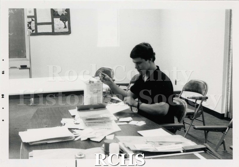 Black and white photograph of the Janesville Public Library Centennial Exhibit Installation. Shows Greg Hartung sitting at a desk with papers spread out on it.