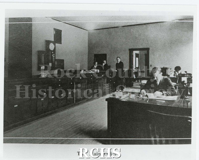 Black and white photograph of the Janesville High School main office. Three standing males. One male and one female seated at desks.