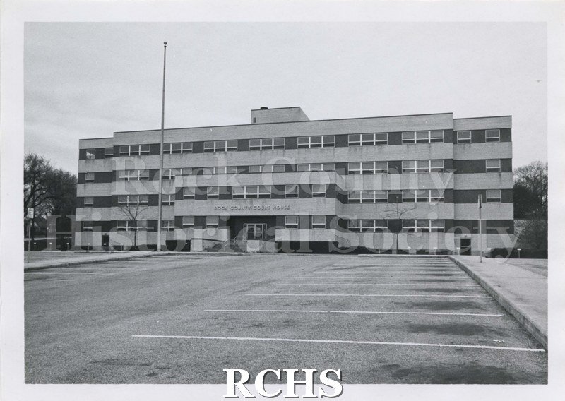 Black and white photograph of Rock County Court House from the front. Shows the parking lot. After image of the Rock County Courthouse-originally shot by John M.