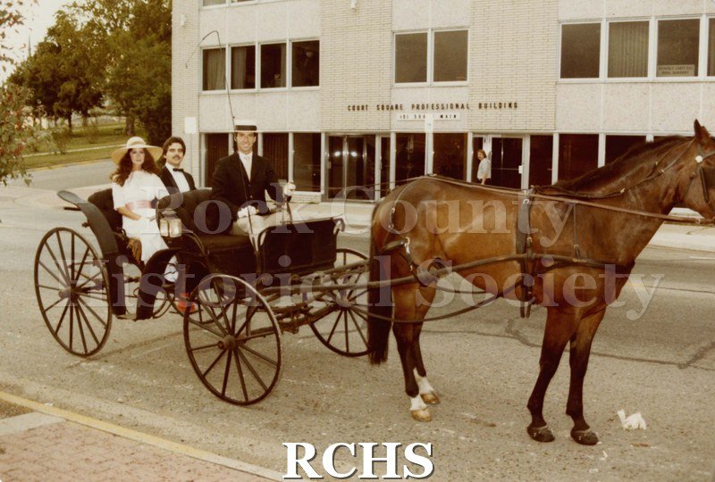 Colored photograph of the Janesville Public Library Centennial Gala . Shows three people seated in an open carriage pulled by one horse.