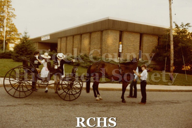 Colored photograph of the Janesville Public Library Centennial Gala. Shows three people dismounting from the open carriage in front of the library.