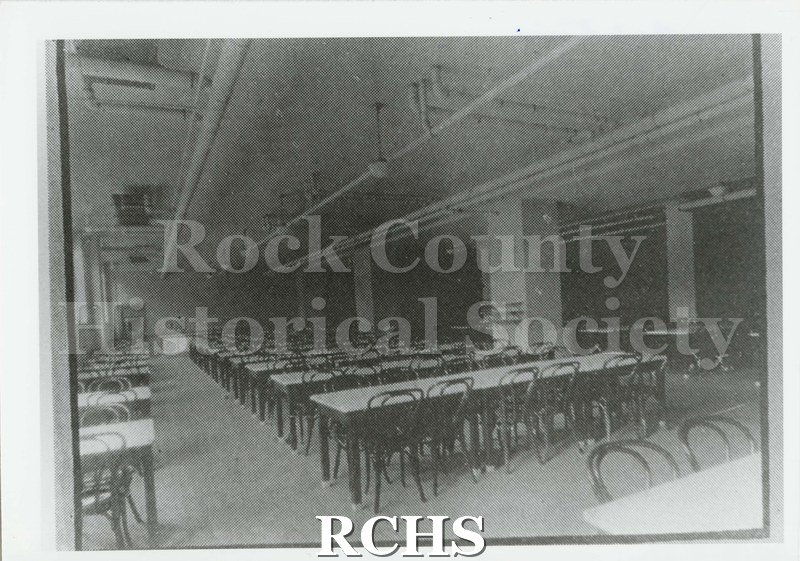 Black and white photograph of the Janesville High School cafeteria. Several long, rectangular tables and chairs. Written on reverse: "Cafeteria 1924 Phoenix pg. 19".