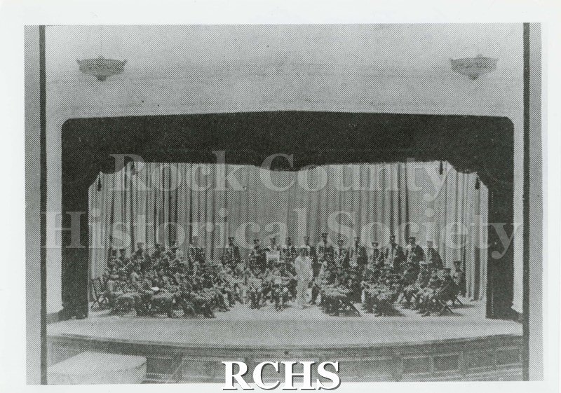 Black and white photograph of the Janesville high school auditorium stage with seated and standing musicians.