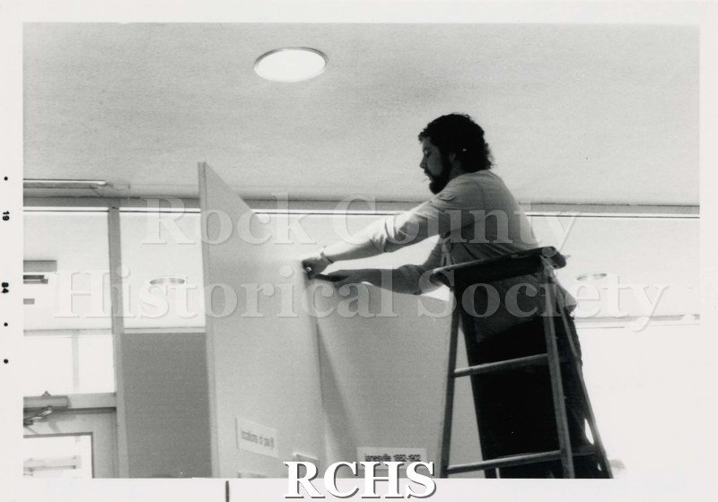 Black and white photograph of the Janesville Public Library Centennial Exhibit Installation showing a man putting up an exhibit. He is standing on a ladder.