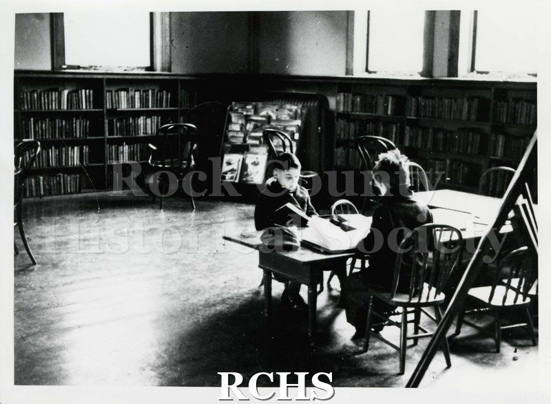 Black and white photograph of two unidentified boys reading books while seated at a table in the library.