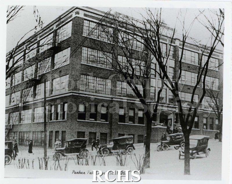 Black and white photograph of the Parker Pen Company circa 1920, taken after a snowfall. Five vehicles are parked on street in front of building.