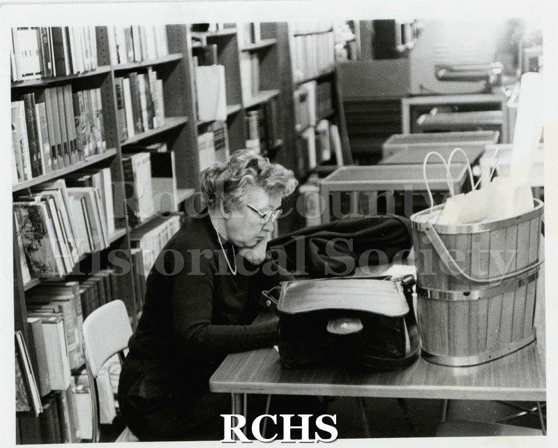 Black and white photograph of Virginia Bell sitting at table in front of bookstack. Written on reverse: "Virginia (Mrs. Marshall) Bell. c.1983 JHS Project Director".