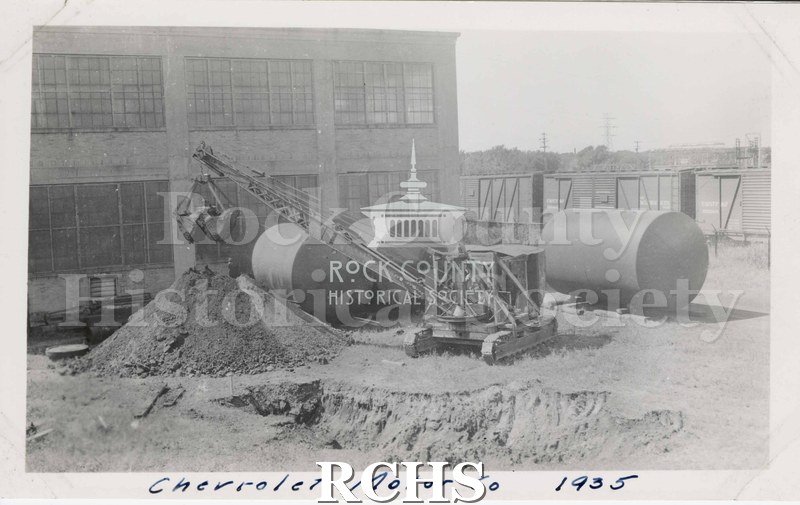 Black and white photograph of Chevrolet Motor Co. 1935. Shows a building with three large round tanks in front of it. There is a crane in front of a pile of dirt.