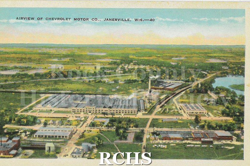 Postcard. Color postcard featuring an aerial view of the Chevrolet factory. Image shows several large factory buildings, railraod tracks, and surrounding fields.
