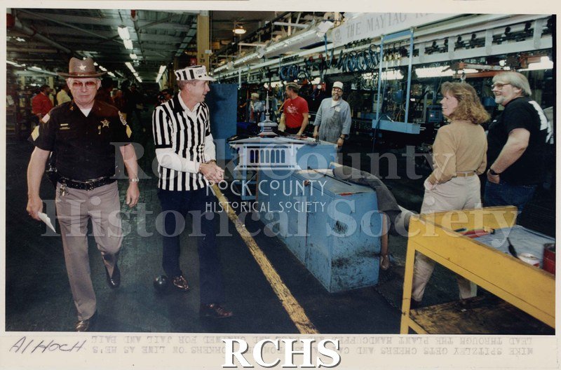 Colored photograph of "Jail and Bail" fund raiser. Shows Sheriff's deputy walking beside Mike Spitzley while workers are standing near him.