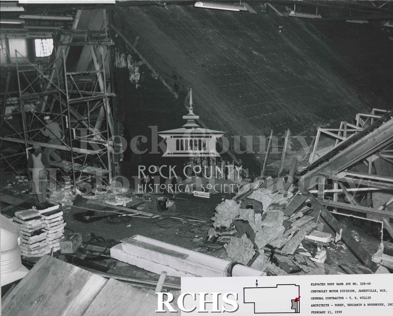 Black and white photograph of the inside the Chevrolet factory. Shown is a lot of debris on the right side and two construction workers and the left side.