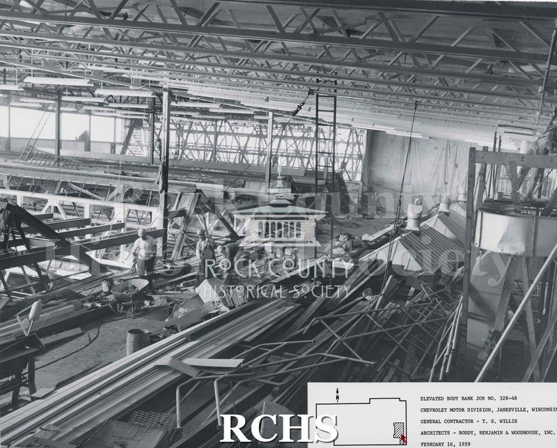 Black and white photograph of the nside of the Chevrolet factory. There are three workmen in the middle of the area, along with a wheelbarrow, wooden barrel and.