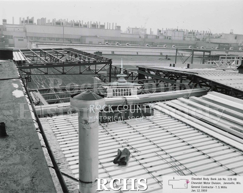 Black and white photograph of the roof of the Chevrolet building. There is a vent pipe in the front with a tarp sitting near it. The factory is in the background.