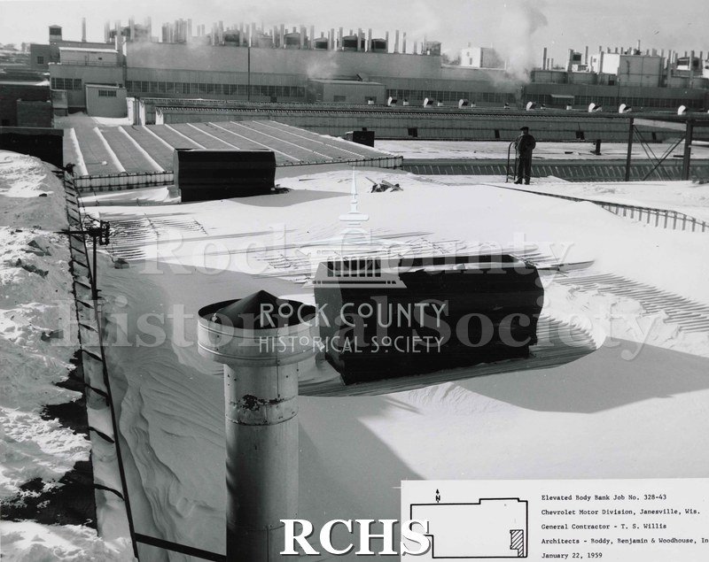 Black and white photograph of the roof of the Chevrolet building. There is a man standing in the snow looking toward the Chevrolet plant.