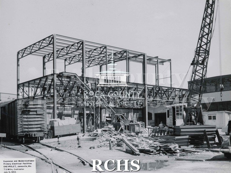 Black and white photograph of a part of the Chevrolet factory construction. Shows a railroad car on the tracks on the left and construction workers.