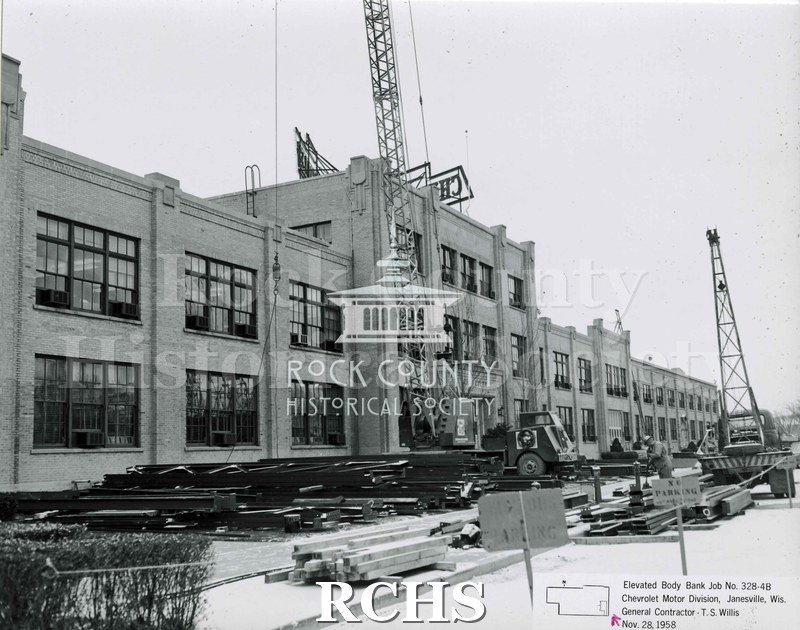 Black and white photograph of the front of the Chevrolet factory. There is a crane in front of the building and another mounted on a truck on the right side.