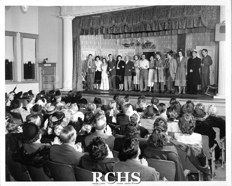 Black and white photograph of fourteen actors taking a curtain call in front of an audience. The set is the same as in 1987.61.24. The house is full.