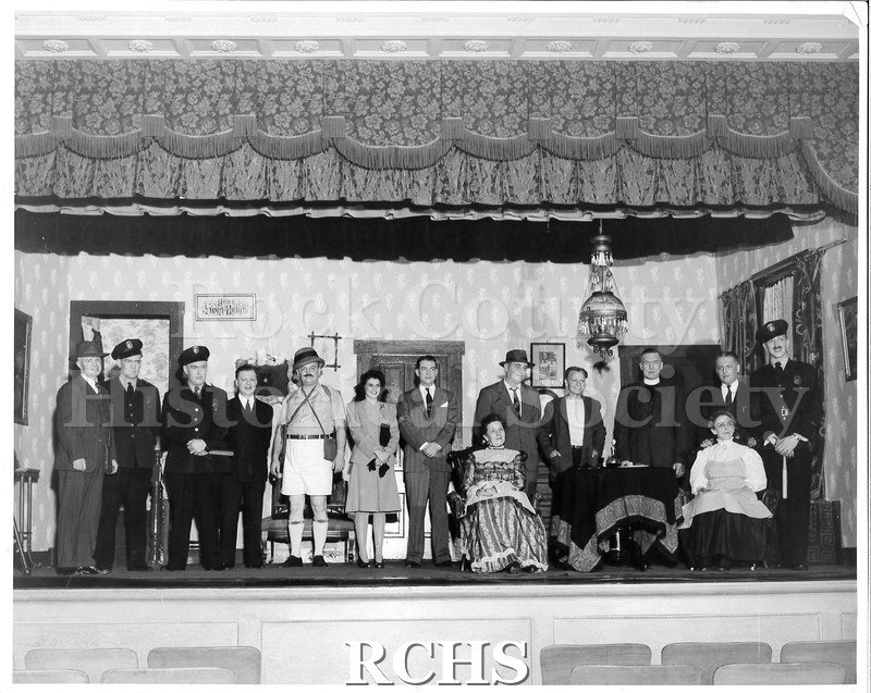 Black and white photograph of fifteen actors posed onstage in a line. Most are standing, two are seated. The set is a house interior.